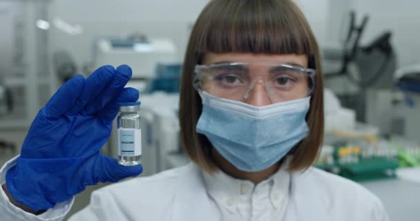 Portrait de femme scientifique en masque de protection et googles regardant à la caméra. Femme en manteau blanc et gants en latex soulevant et tenant le flacon avec le vaccin tout en restant debout en laboratoire. — Video