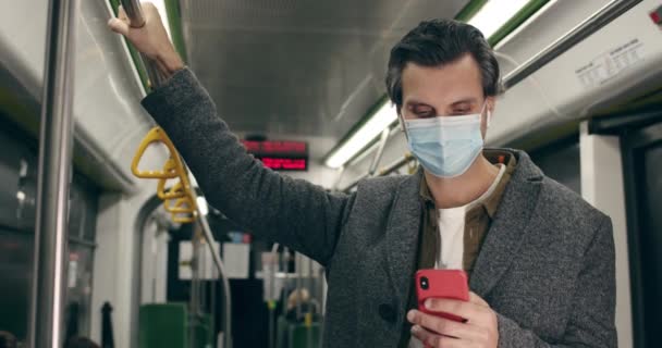 戴着防护口罩的时髦男人用智能手机。在公共交通工具上，男性在无线耳塞中一边聊天一边打字。大流行病的概念. — 图库视频影像