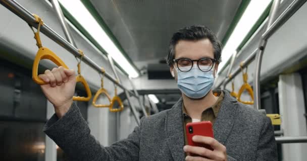 Portrét pohledného muže v lékařské masce a brýlích pomocí chytrého telefonu. Muž ve sluchátkách zvedá hlavua dívá se do kamery při prohlížení interiéru ve veřejné dopravě. — Stock video