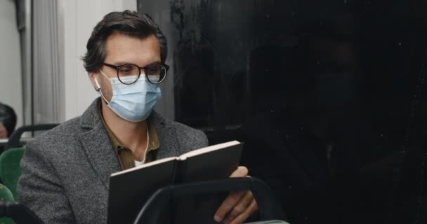 トラムに座っている間、医学マスクの男のミステリービューは本を保持し、咳。公共交通機関で物語を読んでいる間、無線イヤホンの男性は病気を探しています。パンデミックの概念. — ストック動画