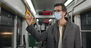 Tramvayda öksüren tıbbi koruyucu maskeli adamın görüntüsü. Gözlüklü ve kablosuz kulaklıklı bir erkek toplu taşımada tırabzan tutarken kendini kötü hissediyor. Salgın kavramı.