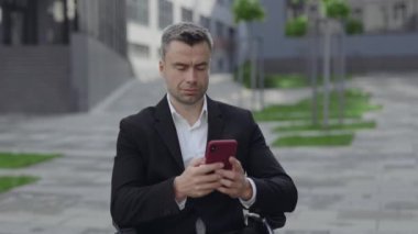 Tekerlekli sandalyedeki başarılı adamın kafasını kaldırmasını ve sokakta internete bakarken kameraya bakmasını yakından izleyin. Takım elbiseli engelli işadamı akıllı telefon kullanıyor ve dışarıda gülümsüyor..
