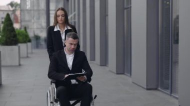Güzel genç bir kadın tekerlekli sandalye itiyor ve engelli patronuyla ofis binasının yakınında konuşuyor. Erkek CEO kadın sekreterle iletişim kurarken belgeleri okuyor..