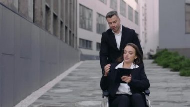 Engelli meslektaşıyla tekerlekli sandalye iten ve iş dünyasıyla iletişim kuran bir adamın görüntüsü. Hasta kadın tablet kullanıyor ve ona yardım eden erkeğe talimat veriyor..
