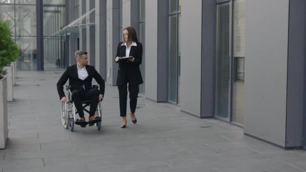 Behinderte männliche Führungskraft im Rollstuhl und Sekretärin bei der Arbeit mit Dokumenten in der Nähe von Bürogebäuden. Mitarbeiter unterhalten sich beim Projektgespräch und gehen auf die Straße. — Stockvideo