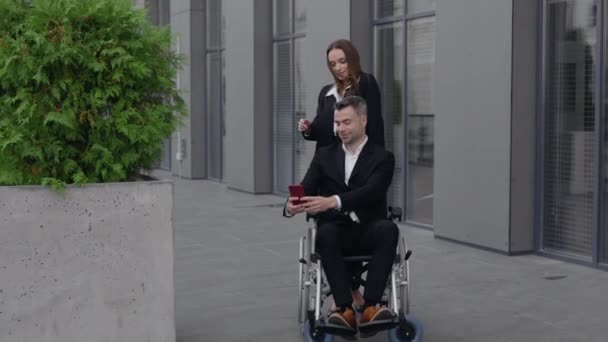 Веселая женщина и мужчина в инвалидной коляске позируют на камеру смартфона, стоя рядом с офисным зданием. Счастливые коллеги-мужчины и женщины делают селфи и улыбаются на улице. Увеличь. — стоковое видео