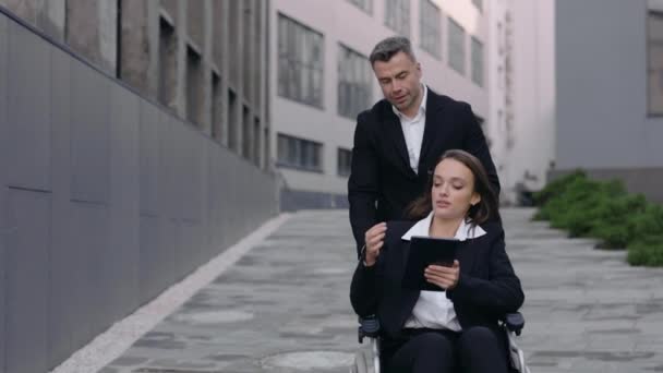 作物的观点，男人推轮椅与他的残疾同事和沟通在西班牙。使用平板电脑的女性残疾人，并向帮助她的男性指明方向. — 图库视频影像