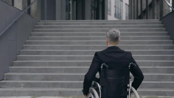 Vista trasera de hombre en traje en silla de ruedas de pie en la calle. Hombre discapacitado parando cerca de las escaleras del edificio de oficinas. Concepto de obstáculos, barreras, indefensos. Acercar. — Vídeos de Stock