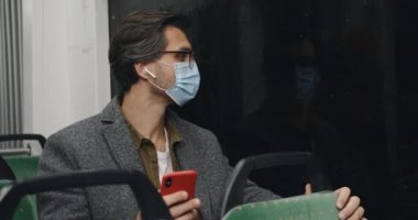 Medikal maskeli, gözlüklü, akıllı telefon kullanan bir adamın görüntüsü. Kulaklıklı bir erkek pencereye bakarken ve akşam toplu taşıma araçlarında otururken telefon ekranına dokunuyor..