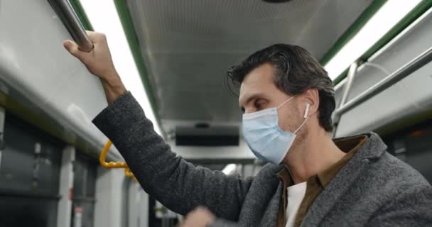 Вид сбоку на парня в медицинской маске, страдающего кашлем и больного в автобусе. Мужчина в беспроводных наушниках плохо себя чувствует и держит перила в общественном транспорте. Понятие пандемии, болезни. — стоковое видео
