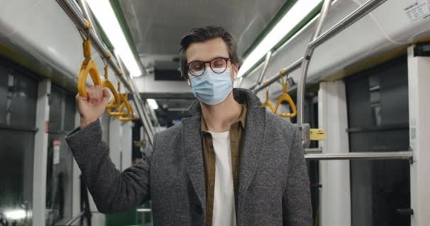 戴着防护性医疗面罩的男人的画像，看着相机。乘坐公共交通工具时，手持扶手的无线耳塞的男性。大流行病概念、检疫. — 图库视频影像