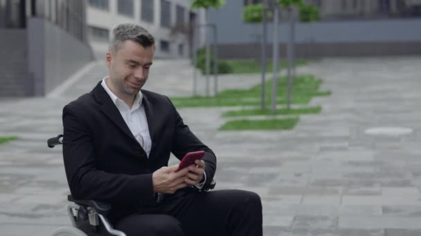 Resmi takım elbiseli mutlu bir adamın şehir caddesinde tekerlekli sandalyede otururken akıllı telefon kullanması ve sevinmesi. Engelli işadamı gülümsüyor ve ofis binasının yakınında evet hareketi yapıyor.. — Stok video
