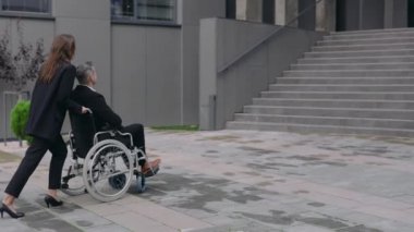 Takım elbiseli şık bir kadın, erkek iş arkadaşıyla tekerlekli sandalye itiyor ve ofis merdivenlerinden önce duruyor. Kadın ve engelli erkek iş adamları işe koyuluyor. Destek ve yardım kavramı.