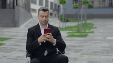 Şehir caddesinde tekerlekli sandalyede otururken akıllı telefon kullanan resmi takım elbiseli neşeli adam manzarası. Ofis binasının yakınında sohbet ederken gülümseyen ve daktilo kullanan engelli işadamı.