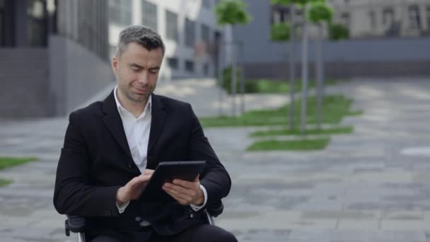 Crop view of cheerful man in wheel chair scrolling, and touching screen while using tablet at city street. Disabled successful businessman in suit browsing internet and smiling. — Stock Video