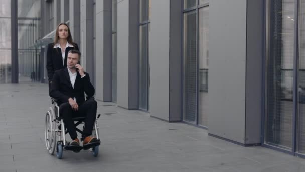 漂亮的年轻女子和她的残疾老板一起推着轮椅在办公大楼附近行走。男首席执行官和他的女秘书一起在智能手机上交谈时提供帮助. — 图库视频影像