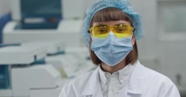 Koruyucu maskeli ve sarı Google 'da kafasını çevirip kameraya bakan bir kadın. Laboratuvarda beyaz önlüklü ve kabarık şapkalı kadın araştırmacı portresi. Tıp kavramı.