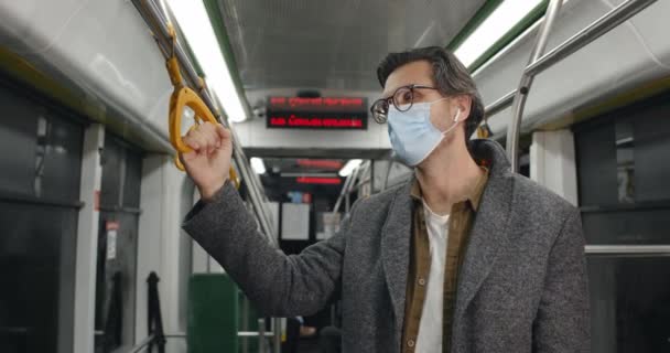 Crop view of man in medical protective mask toux and looking bad in bus. Homme en lunettes et écouteurs sans fil se sentant malade tout en tenant la main courante dans les transports en commun. — Video