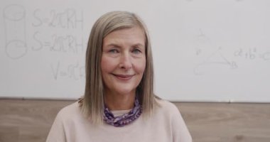 Kameraya bakarken gülümseyen olgun bir kadının portresi. Sınıfta oturan neşeli bayan profesörü yakından çek. Çevrimiçi ve uzak öğrenim kavramı.