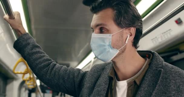 Крупный план красивого мужчины в защитной медицинской маске с помощью смартфона. Мужчина в беспроводных наушниках слушает музыку и держит перила под землей. Концепция пандемии. — стоковое видео