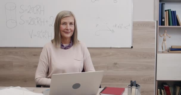 Porträt einer fröhlichen Frau, die in die Kamera blickt. Großaufnahme einer Lehrerin, die am Schreibtisch vor dem Laptop im Klassenzimmer sitzt und lächelt. Konzept des Online-Lernens. — Stockvideo