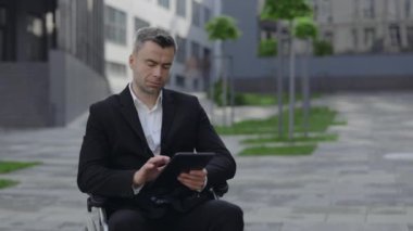 Şehir caddesinde tekerlekli sandalyede otururken resmi kıyafetli bir adamın tablet kullanması. Çalışmayan başarılı işadamı ofis binası yakınlarında e- posta yazarken aygıt ekranına dokunuyor.
