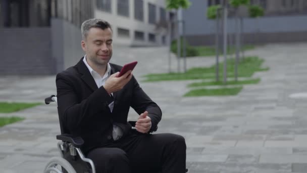 Oříznout pohled na pozitivního zdravotně postiženého muže ve formálním obleku, který diktuje hlasovou zprávu a posílá ven. Úspěšný podnikatel v invalidním vozíku mluvit a používat smartphone mikrofon při úsměvu. — Stock video