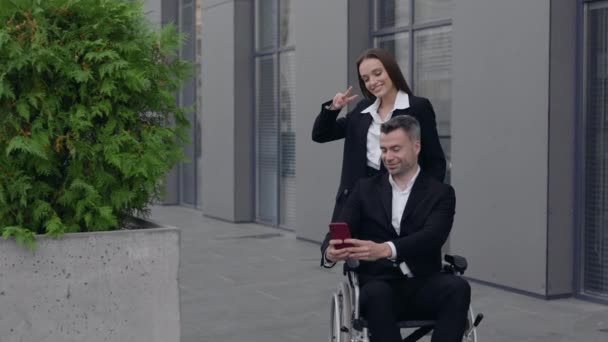 Kvinna och man i rullstol poserar till smartphone kamera när du står nära kontorsbyggnad. Manliga och kvinnliga medarbetare ler medan de gör selfies och tittar på skärmen. Zooma in. — Stockvideo