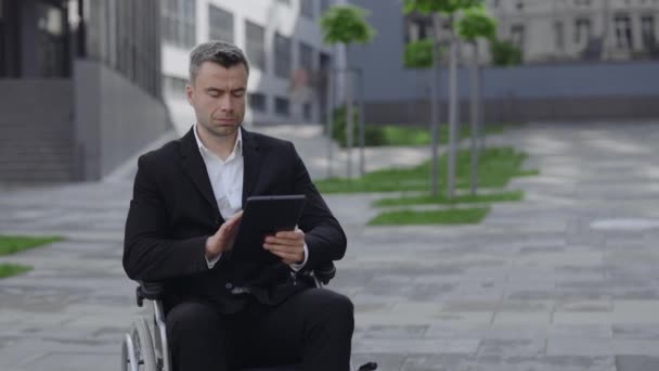 Vista de la cosecha de hombre guapo en silla de ruedas desplazamiento, y la pantalla táctil mientras se utiliza la tableta en la calle de la ciudad. Discapacitados empresario concentrado en traje de navegación por Internet cerca de la oficina. — Vídeos de Stock