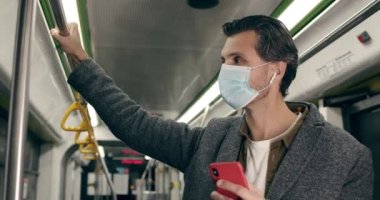 Akıllı telefon kullanan koruyucu tıbbi maskeli bir adamın görüntüsü. Kablosuz kulaklıklı, müzik dinleyen ve toplu taşımada tırabzan tutan bir erkek. Salgın kavramı.
