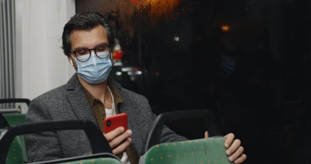 Oříznout pohled na člověka v ochranné masce a brýlích pomocí chytrého telefonu. Muž pohledný člověk ve sluchátkách procházející internet při pohledu na okno a sedí v tramvaji ve večerních hodinách. — Stock video
