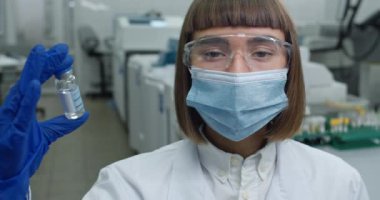 Koruyucu maskeli kadın bilim adamı ve Google 'da kafasını çevirip kameraya bakıyor. Beyaz önlüklü ve lateks eldivenli kadın laboratuarda dururken cam şişeyi aşıyla gösteriyor..