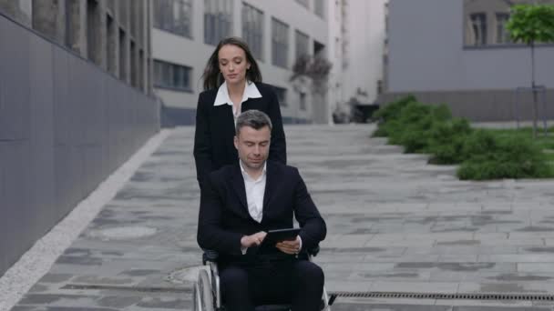 Widok plonu młodej kobiety pchającej wózek inwalidzki ze swoim niepełnosprawnym szefem podczas spaceru w pobliżu biurowca. mężczyzna biznesmen za pomocą tablet a idąc z jego kobieta sekretarz pomoc. — Wideo stockowe