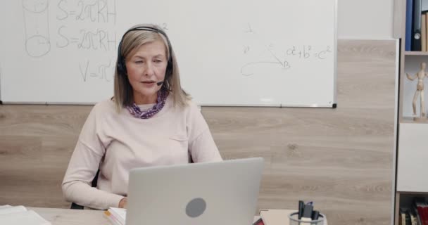 무선 헤드셋을 쓴 긍정적 인 여성 교사가 노트북 화면을 보면서 마이크로 대화하고 손을 흔들며 작별 인사를 했습니다. 온라인 수학 수업을 받는 여성. 거리 학습에 대한 개념. — 비디오