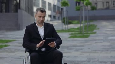 Tekerlekli sandalyedeki yakışıklı adamın kafasını kaldırmasını ve caddede kameraya bakmasını yakından izleyin. Tablet kullanan ve dışarıda gülümseyen takım elbiseli başarılı bir iş adamı. Yakınlaştır.