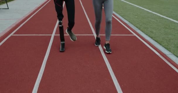 Crop view of disabled man with amputated leg and sports woman jogging. Male sportsman with running blade and female person run on race track at stadium. Concept of motiation, health, sports. — Wideo stockowe