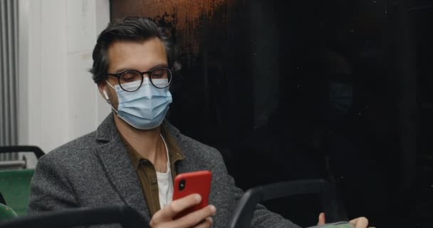 Male person in wireless earbuds listening to music and touching screen while sitting in public transport. Guy in medical mask and glasses using smartphone. Concept of pandemic, commuting. — Stok video