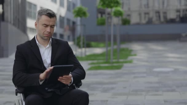 Crop view of serious man in wheel chair scrolling, zooming and touching screen while using tablet at city street. Disabled businessman in suit working with documents near office building. — Stock Video