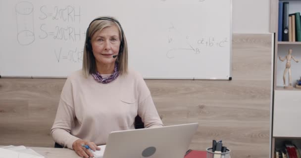 Portret dojrzałej kobiety w słuchawkach bezprzewodowych z mikrofonem patrzącym w kamerę. Zbliżenie nauczycielki siedzącej przy biurku przed laptopem w klasie. Koncepcja uczenia się przez internet. — Wideo stockowe