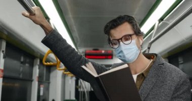 Tıbbi maskeli bir adamın tramvayda kitap tutuşu görüntüsü. Gözlüklü, kablosuz kulaklıklı, hikaye okuyan ve toplu taşımada tırabzan tutan bir erkek. İşe gidip gelme kavramı.