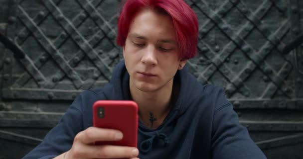 Lwów, Ukraina - 25 lipca 2019: Mężczyzna hipster w ciemnej bluzie przewijanie wiadomości i patrząc na ekranie telefonu. Młody facet z farbowanymi rudymi włosami za pomocą smartfona do przeglądania Internetu podczas siedzenia na zewnątrz. — Wideo stockowe