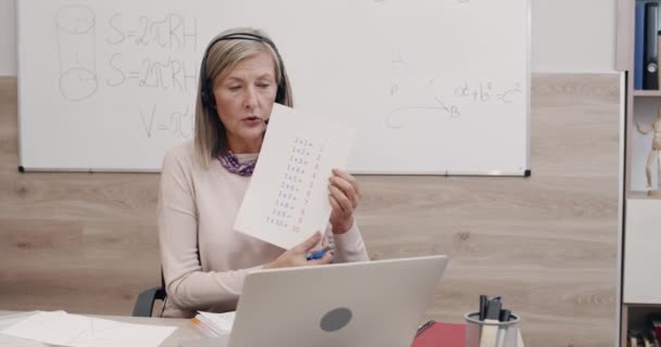 Insegnante femminile prendendo e mostrando tabella di moltiplicazione mentre guardando lo schermo del computer portatile. Donna in cuffia con microfono insegnare matematica mentre seduto alla scrivania. Concetto di apprendimento a distanza. — Video Stock