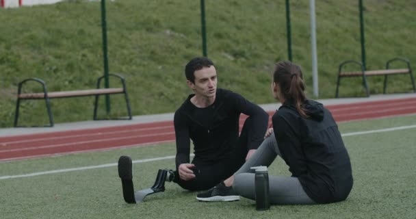 Ein behinderter junger Mann mit einer Prothese im Gespräch mit einer Frau. Männlicher Athlet mit amputiertem Bein und weibliche Person kommunizieren, während sie auf dem Rasen am Sportplatz sitzen. Gesundheitskonzept. — Stockvideo