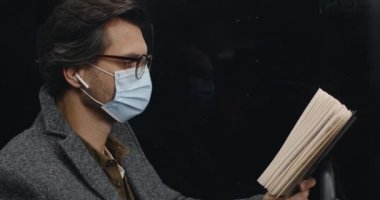 Tramvayda otururken kitap sayfasını çeviren koruyucu maskeli zeki adamın yan görüntüsü. Kablosuz kulaklıklı bir erkek toplu taşıma araçlarında hikaye okuyor. İşe gidip gelme kavramı.