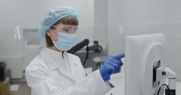 Vista de la cosecha de la trabajadora en máscara protectora y googles introduciendo datos en el ordenador. Científico en rob y tapón bouffant sosteniendo botella médica de vidrio mientras escribe en la pantalla en el laboratorio. — Vídeos de Stock