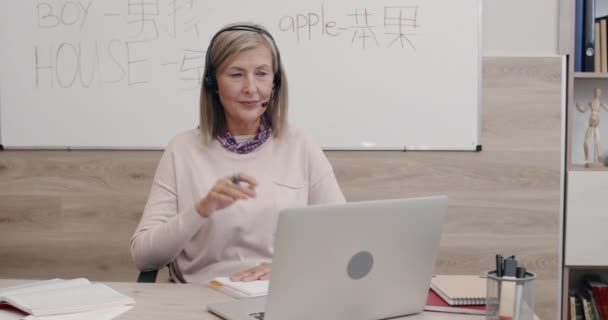 Kulaklıklı, mikrofonlu yaşlı bir kadın. Dil öğretirken bilgisayar ekranına bakıyor. İnternet dersinde masa başında oturan kadın profesör. Uzaklık öğrenme kavramı. — Stok video