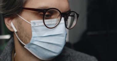 Tıbbi koruyucu maskeli yakışıklı adamın otobüste oturmasını yakından izleyin. Kablosuz kulaklıklı ve gözlüklü bir adam toplu taşımada müzik dinliyor. İşe gidip gelme kavramı.