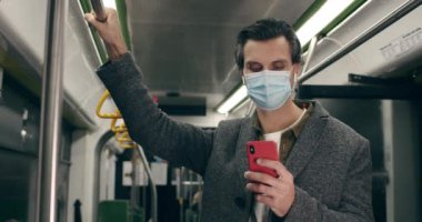 Koruyucu tıbbi maskeli adam akıllı telefon kullanıyor. Toplu taşıma araçlarında dolaşırken kulaklıklı bir erkek telefon ekranına dokunuyor. Salgın, karantina kavramı.