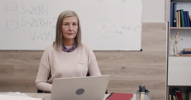 카메라를 보고 있는 쾌활 한 여성 교수의 초상화. 교실에서 책상 앞에 앉아 컴퓨터 앞에서 웃고 있는 여성의 모습을 클로즈업 해 보 세요. 온라인 및 거리 학습의 개념. — 비디오