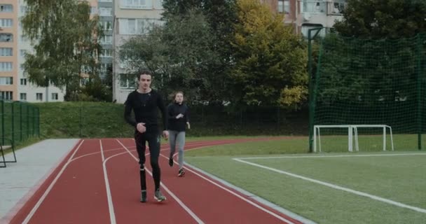 Erős fogyatékos férfi megelőzi a fiatal nőt, miközben versenypályán futnak. Férfi atléta futópengével és női kocogóval a stadionban. A motiváció, az egészség és a sport fogalma. — Stock videók